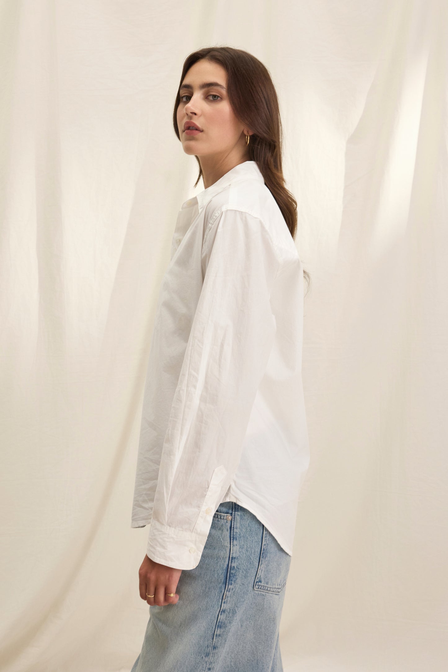 A person in the KELIA SHIRT by Velvet by Graham & Spencer and light blue jeans stands sideways before a light fabric backdrop.-40281806110913
