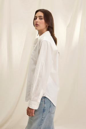 A person in the KELIA SHIRT by Velvet by Graham & Spencer and light blue jeans stands sideways before a light fabric backdrop.