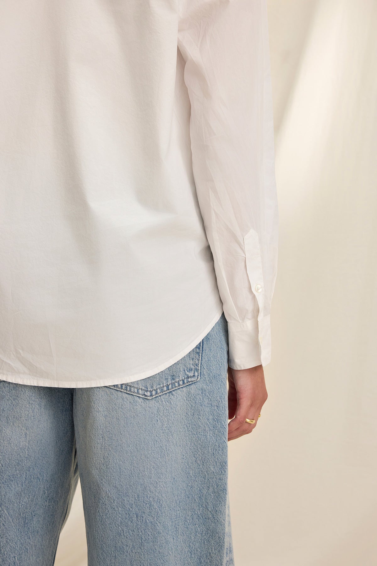 Model in the KELIA SHIRT by Velvet by Graham & Spencer, a white long-sleeve top, paired with light blue jeans, stands against a neutral background with their arm resting by their side.