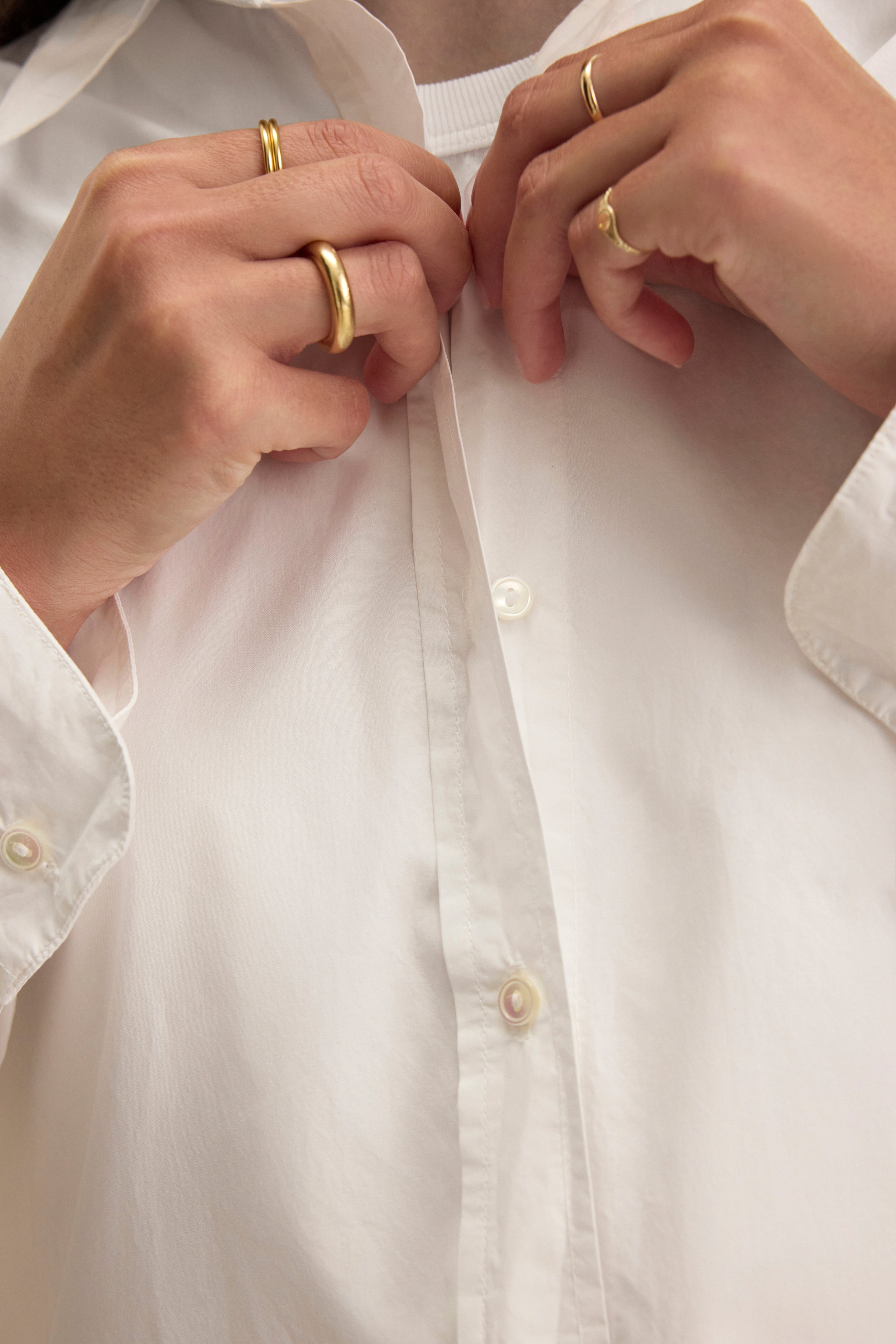 Close-up of a person buttoning the KELIA SHIRT by Velvet by Graham & Spencer, adorned with several gold rings on their fingers.