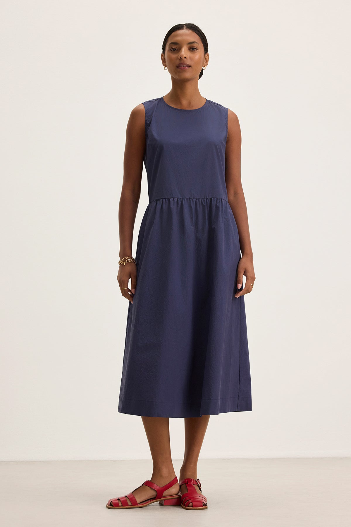 A person in a Velvet by Graham & Spencer MASHA DRESS, featuring a sleeveless navy midi-length design with an elastic waist, and paired with red sandals stands against a plain background.-38662692602049