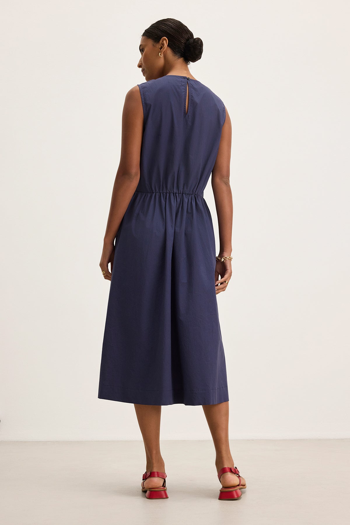 A woman in the Velvet by Graham & Spencer MASHA DRESS, a sleeveless navy cotton poplin midi-length dress, and red sandals stands facing away on a light background.-38662692569281