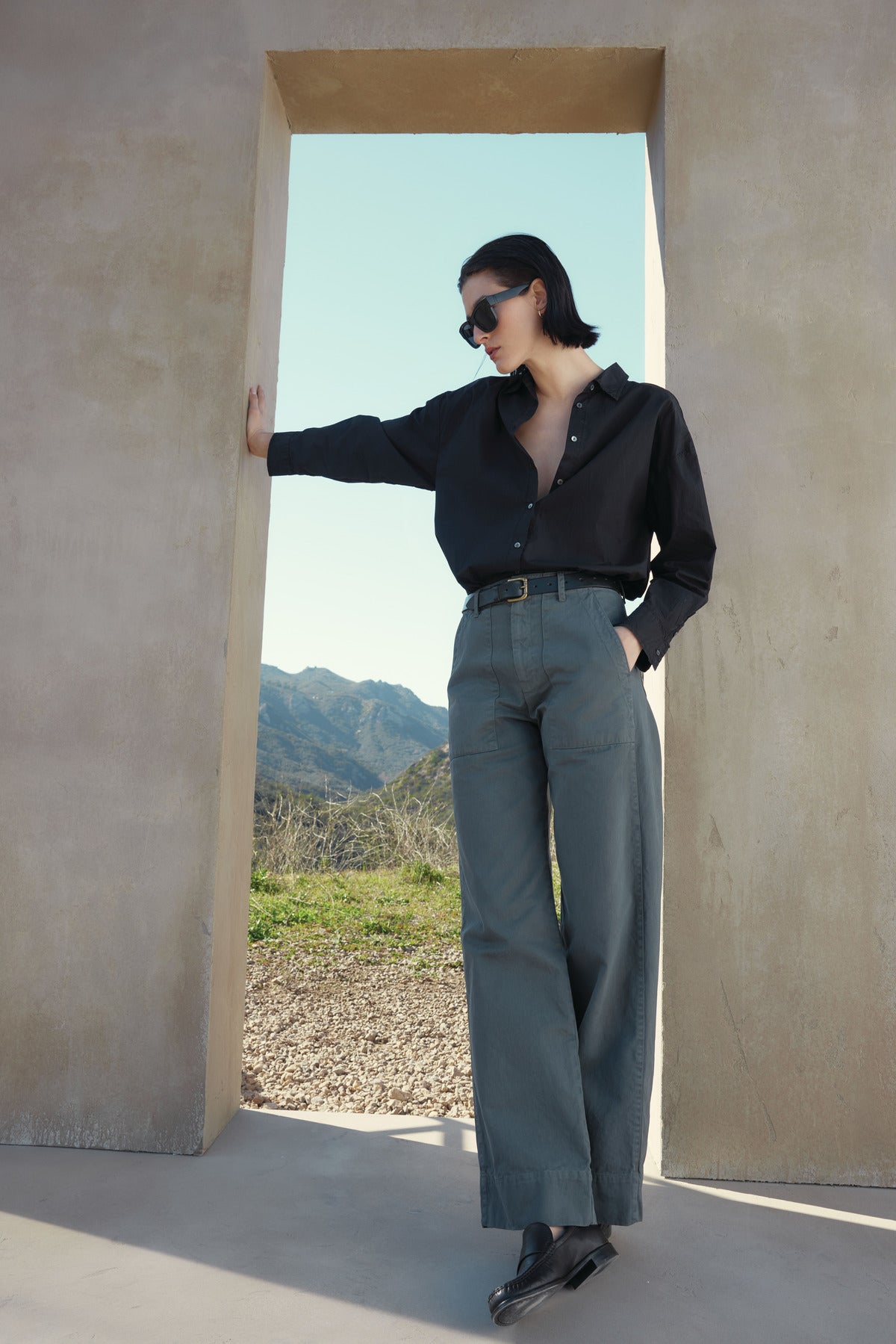 A person in sunglasses, wearing a black DAKOTA SHIRT from Velvet by Jenny Graham and green pants, leans against a stone doorway with a scenic mountain view in the background.-37666924396737