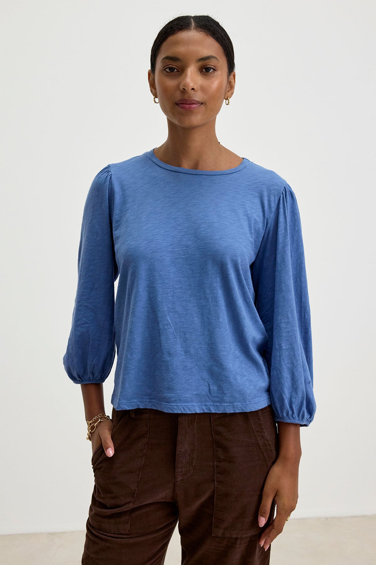   A person is standing against a plain, light background, wearing the ANETTE TEE by Velvet by Graham & Spencer, a versatile blue, long-sleeved shirt made of cotton slub knit, and brown pants. They have their right hand in their pocket and are looking forward. 