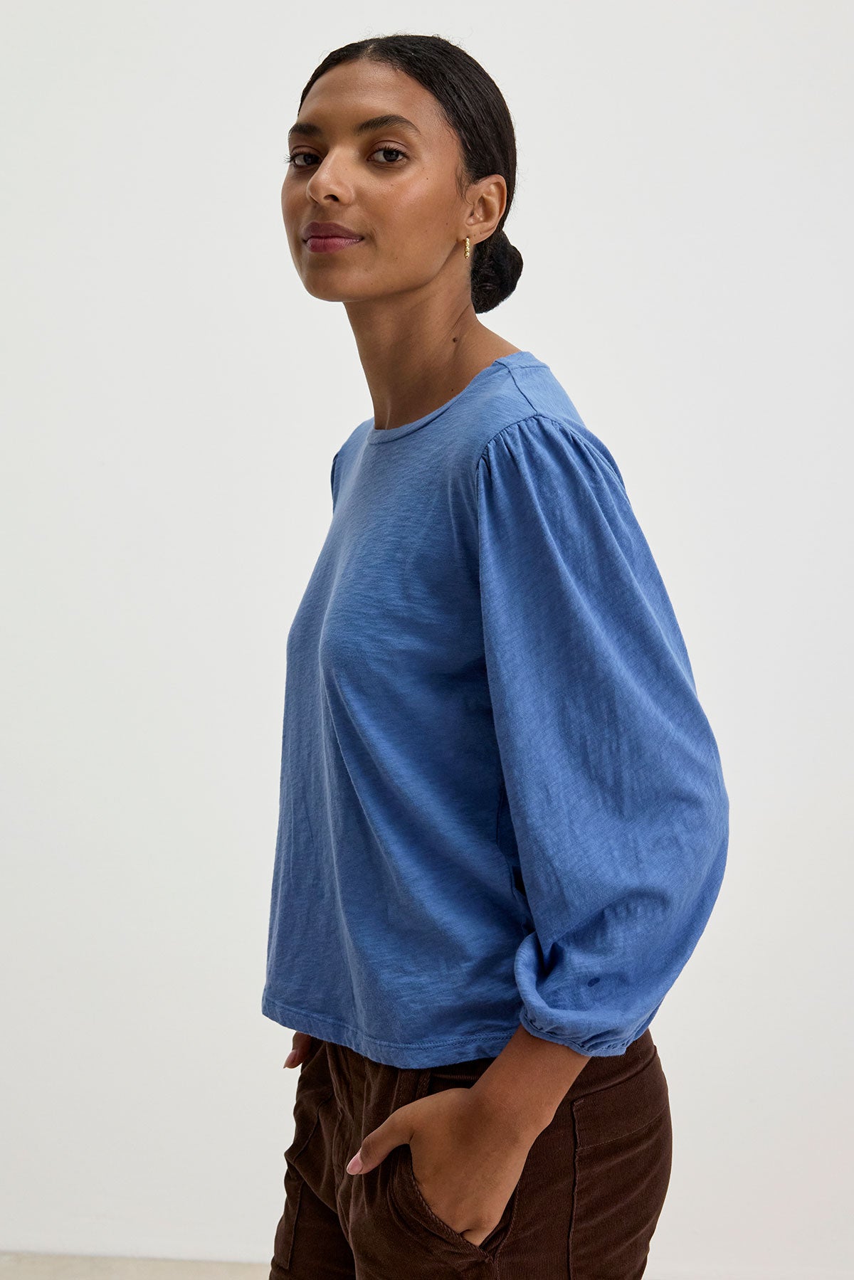   A person stands in a relaxed pose, wearing the ANETTE TEE from Velvet by Graham & Spencer—a versatile blue long-sleeved shirt made from cotton slub knit—and brown pants, against a plain white background. 