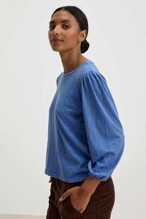 A person stands in a relaxed pose, wearing the ANETTE TEE from Velvet by Graham & Spencer—a versatile blue long-sleeved shirt made from cotton slub knit—and brown pants, against a plain white background.