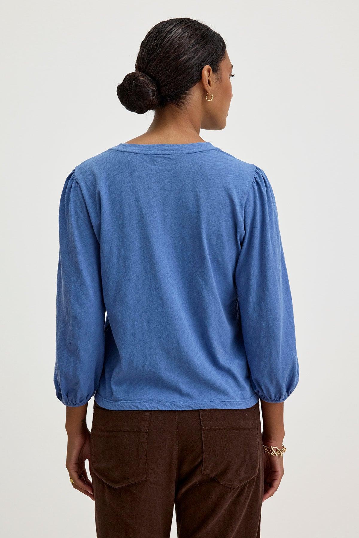   A person with dark hair styled in a bun is wearing the ANETTE TEE by Velvet by Graham & Spencer, a blue top with three-quarter length sleeves made from cotton slub knit, along with brown pants, photographed from the back. 