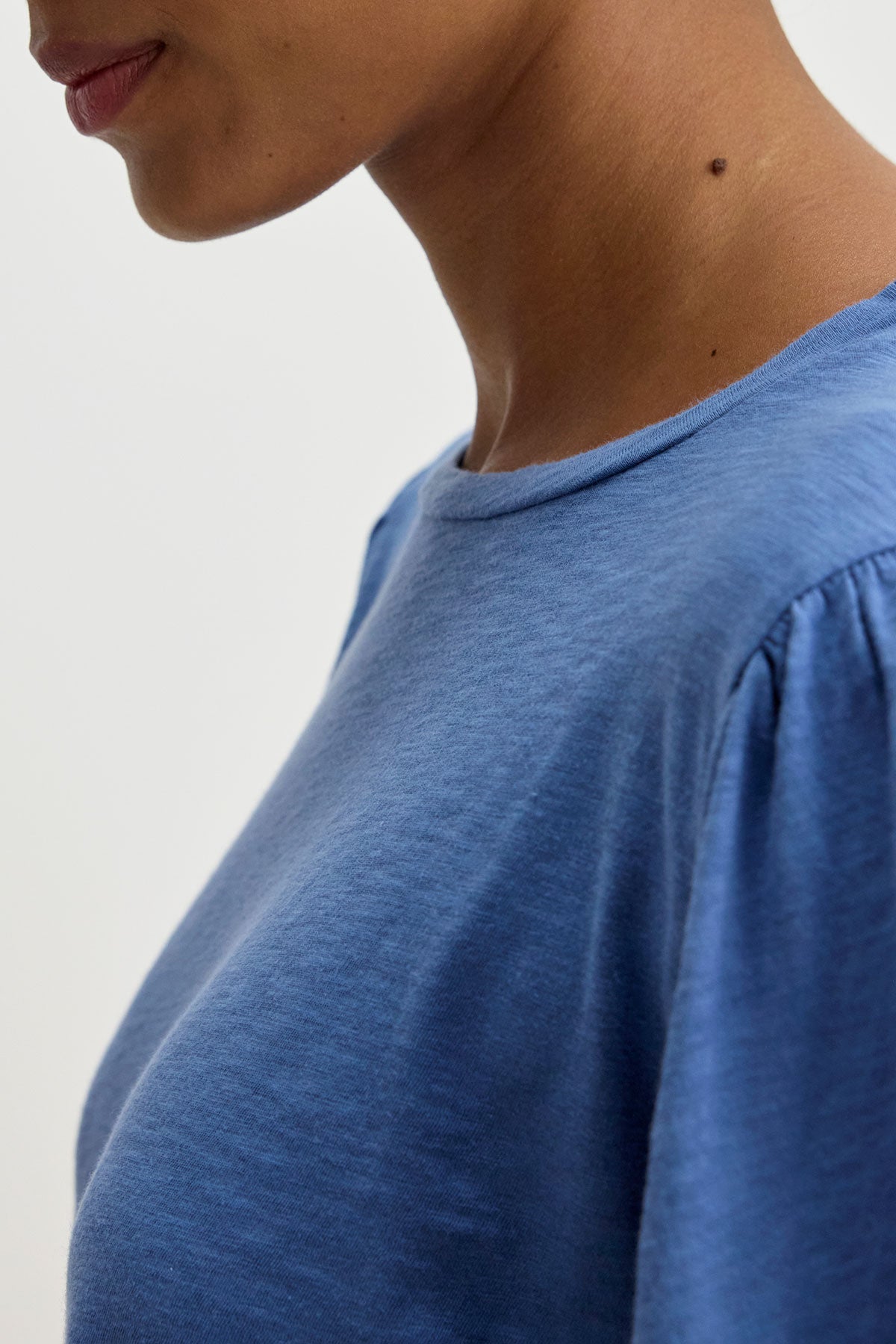   Close-up of a person wearing the ANETTE TEE from Velvet by Graham & Spencer, a versatile blue top made of cotton slub knit, set against a light, neutral background. The focus is on the upper section of the garment and the side of the person's face and neck. 