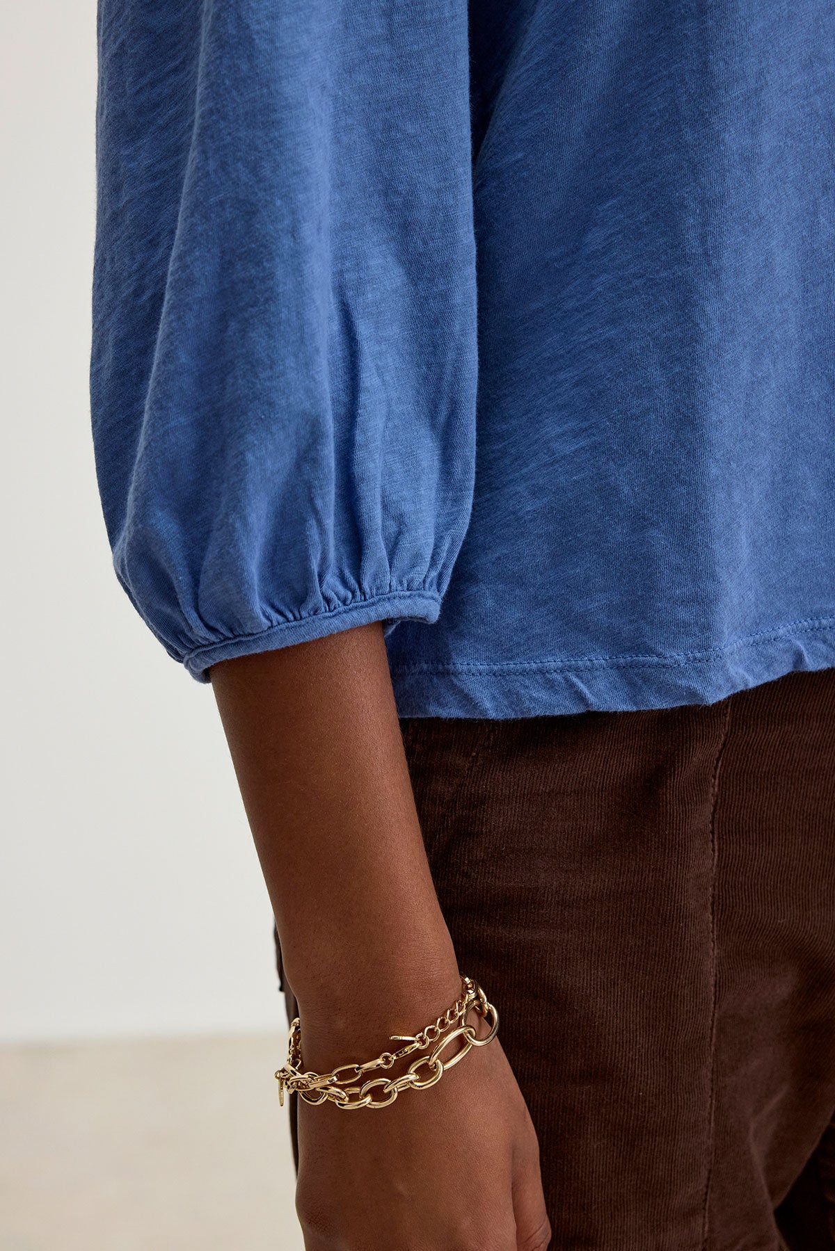   A person wearing a blue ANETTE TEE by Velvet by Graham & Spencer and brown pants with a gold chain bracelet on their wrist. 