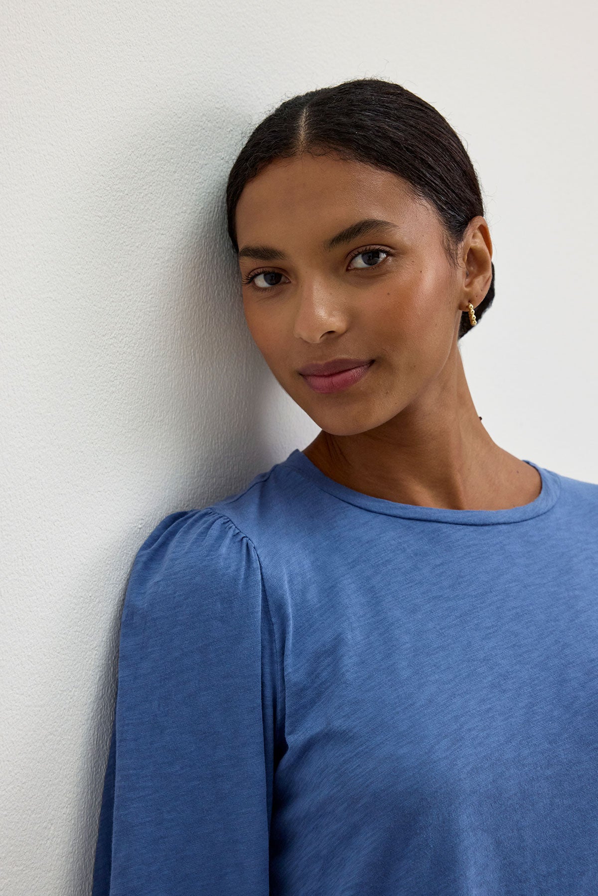 A person with neatly tied hair and wearing an ANETTE TEE by Velvet by Graham & Spencer stands against a white wall, facing the camera with a slight smile.-37745632542913