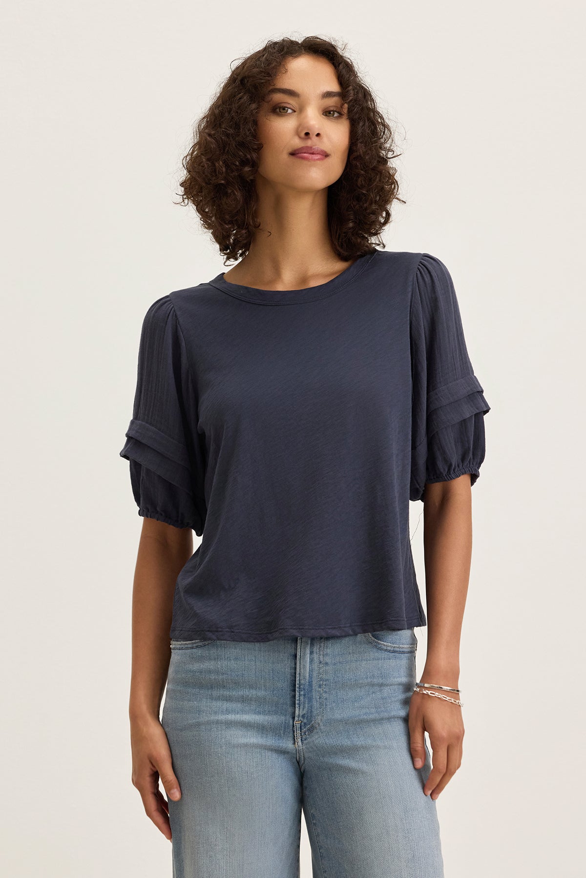 A woman with curly brown hair wears the Velvet by Graham & Spencer HADEN TOP in dark blue with puff sleeves and light blue jeans, standing against a plain light background.-39333200494785