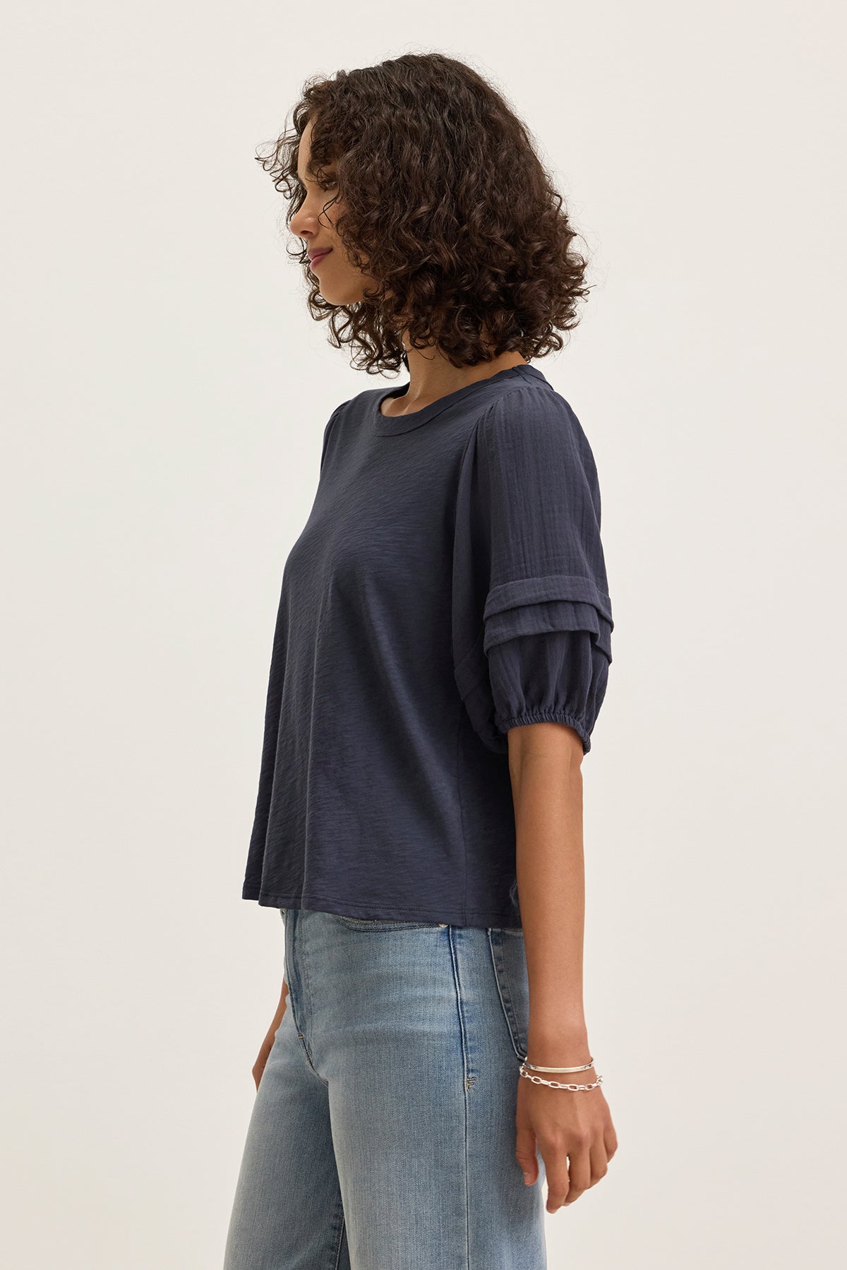A woman stands in profile against a plain background, wearing the HADEN TOP by Velvet by Graham & Spencer—a navy cotton slub short-sleeve top with subtle puff sleeves—paired with light blue jeans.-39333200560321