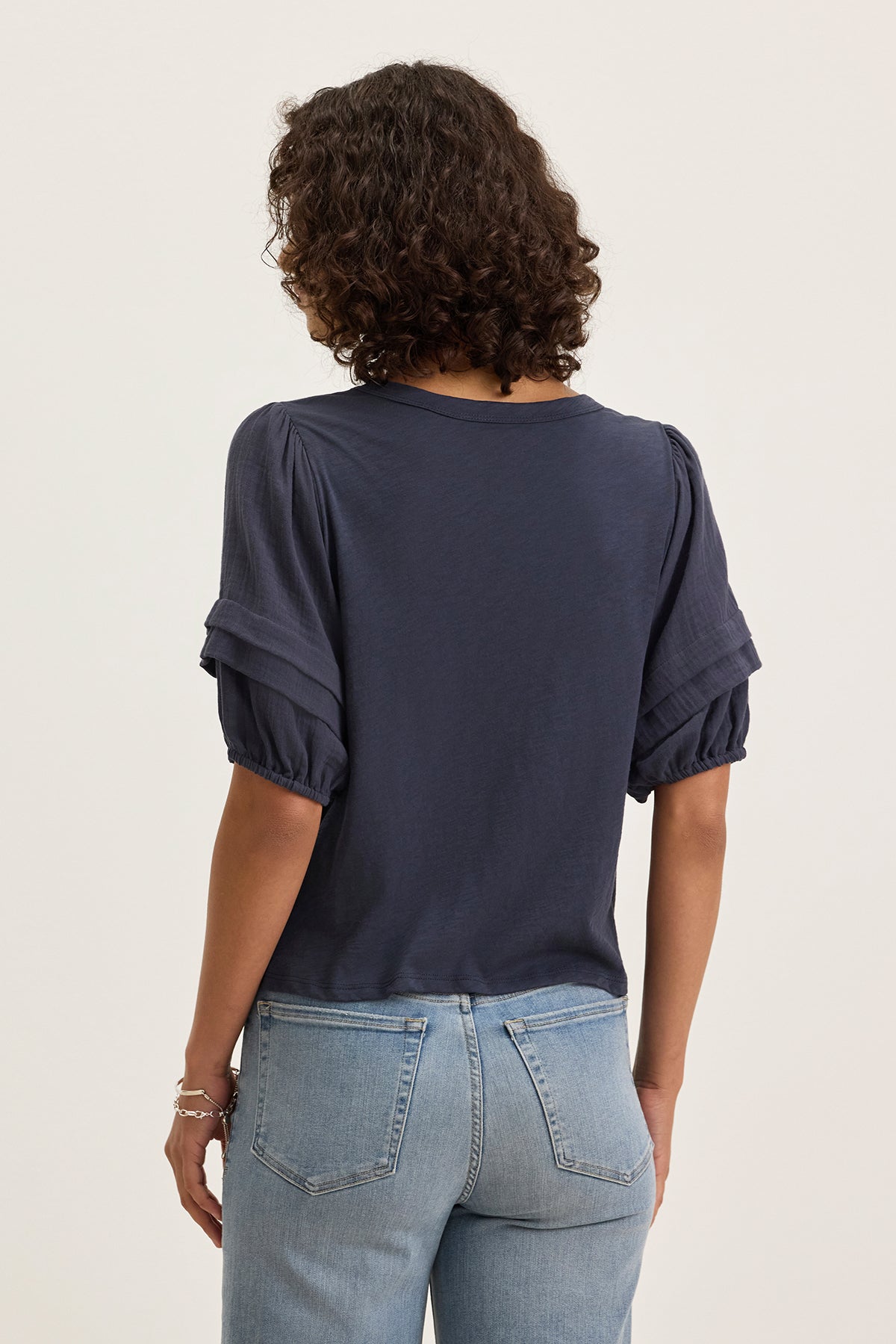 A person with curly hair is seen from the back wearing the HADEN TOP by Velvet by Graham & Spencer, a navy cotton slub short-sleeve top with puff sleeves, paired with light blue jeans against a plain background.-39333200527553