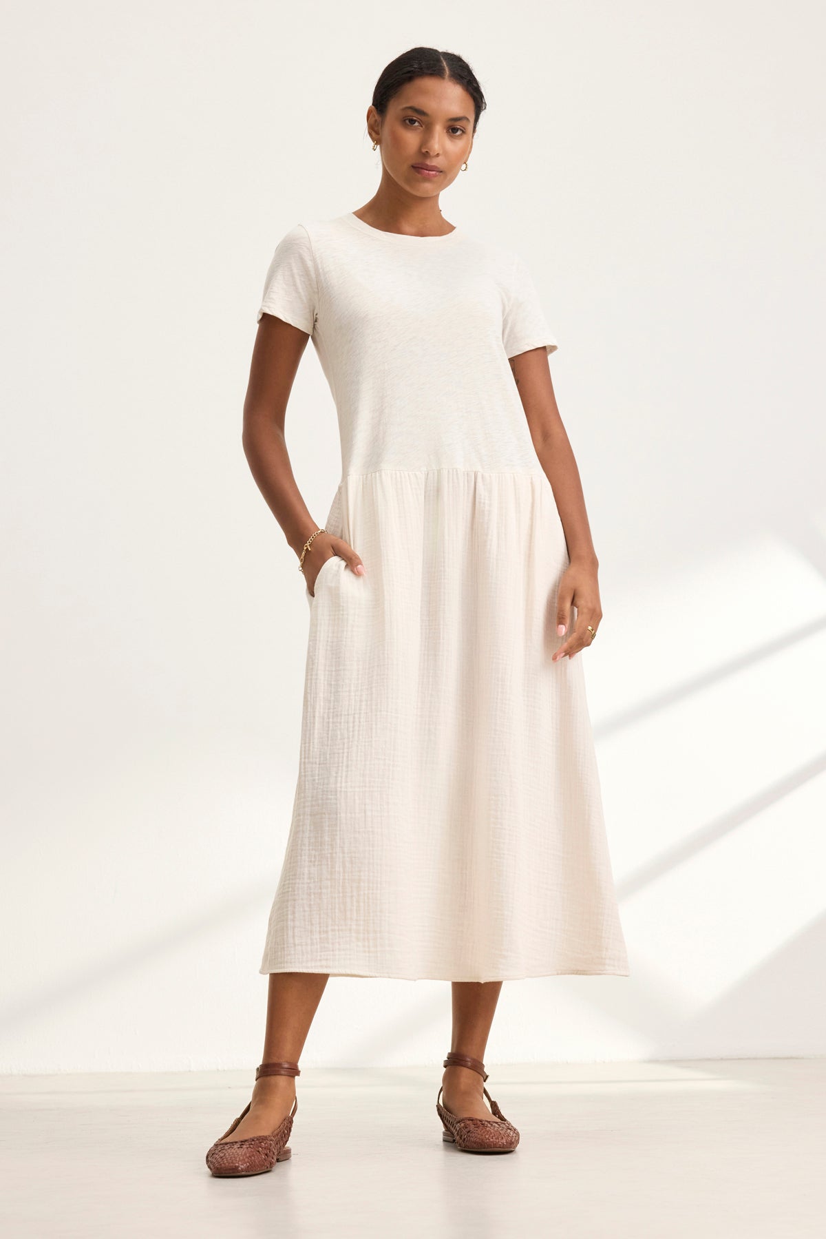   Against a plain white background, a person wears the ITZA DRESS by Velvet by Graham & Spencer, a short-sleeved, cream-colored, ankle-length dress paired with brown sandals. 