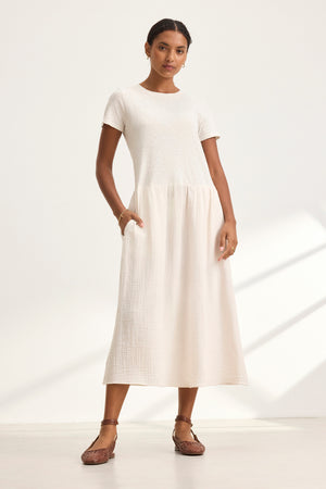 Against a plain white background, a person wears the ITZA DRESS by Velvet by Graham & Spencer, a short-sleeved, cream-colored, ankle-length dress paired with brown sandals.