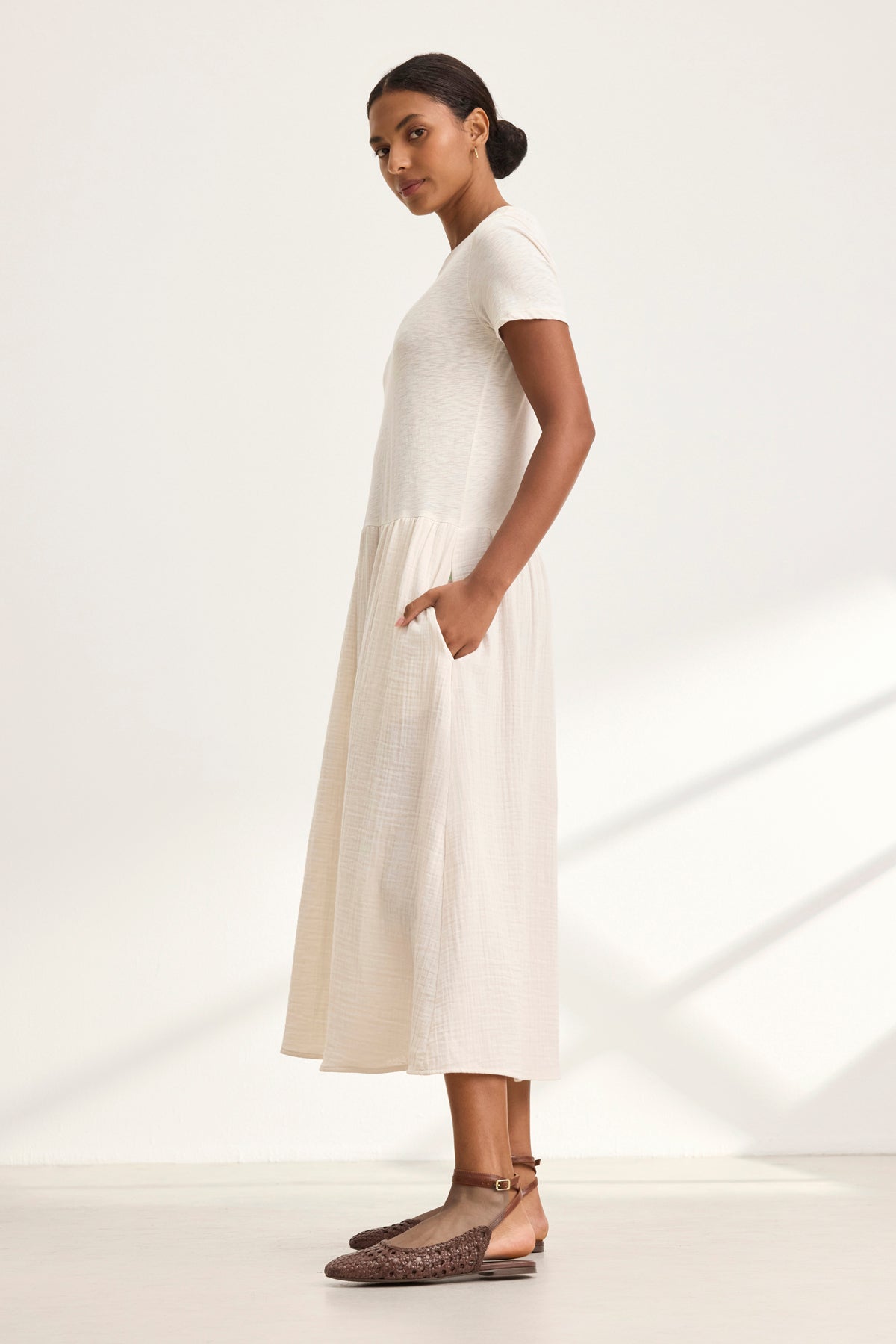   A person poses sideways in a ITZA DRESS by Velvet by Graham & Spencer, featuring a white short-sleeve dress with flared, textured skirt and hands in pockets, paired with brown woven shoes in a simple neutral setting. 