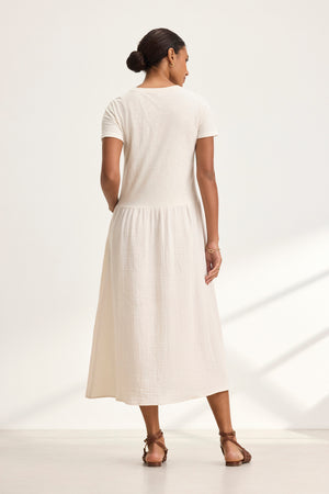 A person wearing the ITZA DRESS by Velvet by Graham & Spencer and sandals stands facing away in a minimalist setting with light gray walls.