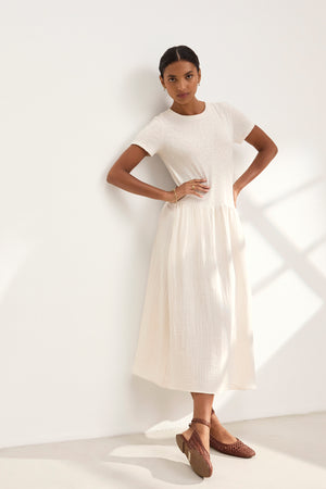 A woman in the ITZA DRESS by Velvet by Graham & Spencer leans against a wall in soft lighting, wearing brown woven shoes.
