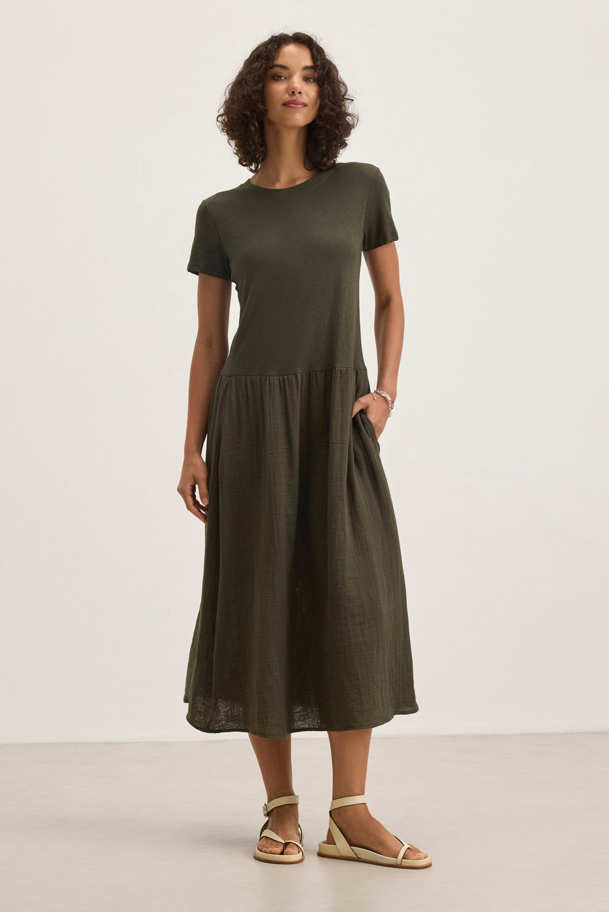 A woman in the Velvet by Graham & Spencer ITZA DRESS, a dark green, short-sleeved midi dress, paired with white sandals, stands on a light floor against a plain background.-38918714392769