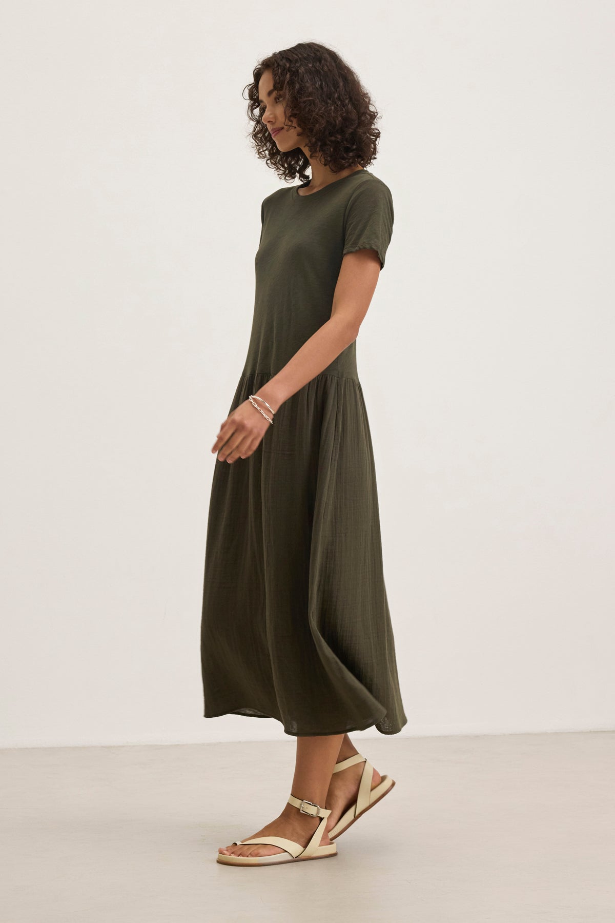   A person with curly hair is wearing the ITZA DRESS by Velvet by Graham & Spencer, featuring a dark green, short-sleeved, ankle-length design and paired with sandals, standing against a plain backdrop. 