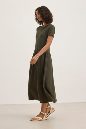 A person with curly hair is wearing the ITZA DRESS by Velvet by Graham & Spencer, featuring a dark green, short-sleeved, ankle-length design and paired with sandals, standing against a plain backdrop.
