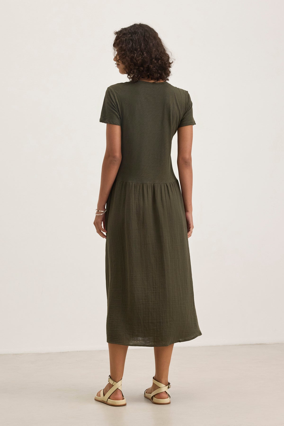 A person faces away against a neutral background, wearing the ITZA DRESS (an olive green short-sleeve midi dress) by Velvet by Graham & Spencer and beige sandals.-38918714327233