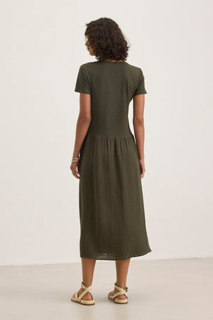 A person faces away against a neutral background, wearing the ITZA DRESS (an olive green short-sleeve midi dress) by Velvet by Graham & Spencer and beige sandals.