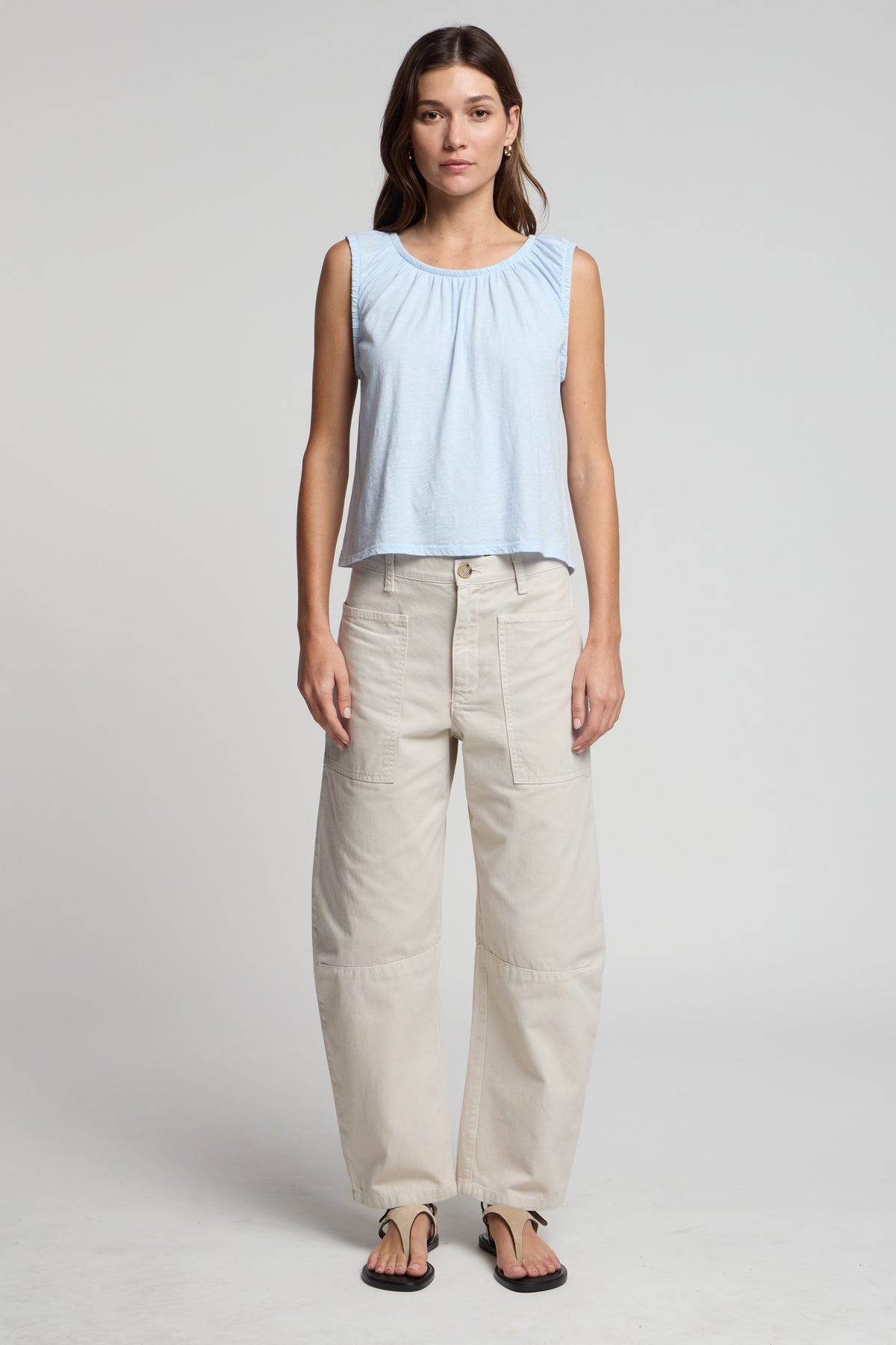 A woman stands facing forward against a plain light background, wearing the JUNA TANK TOP by Velvet by Graham & Spencer—a light blue sleeveless top—paired with beige wide-leg pants with patch pockets and black sandals.-41569250181313
