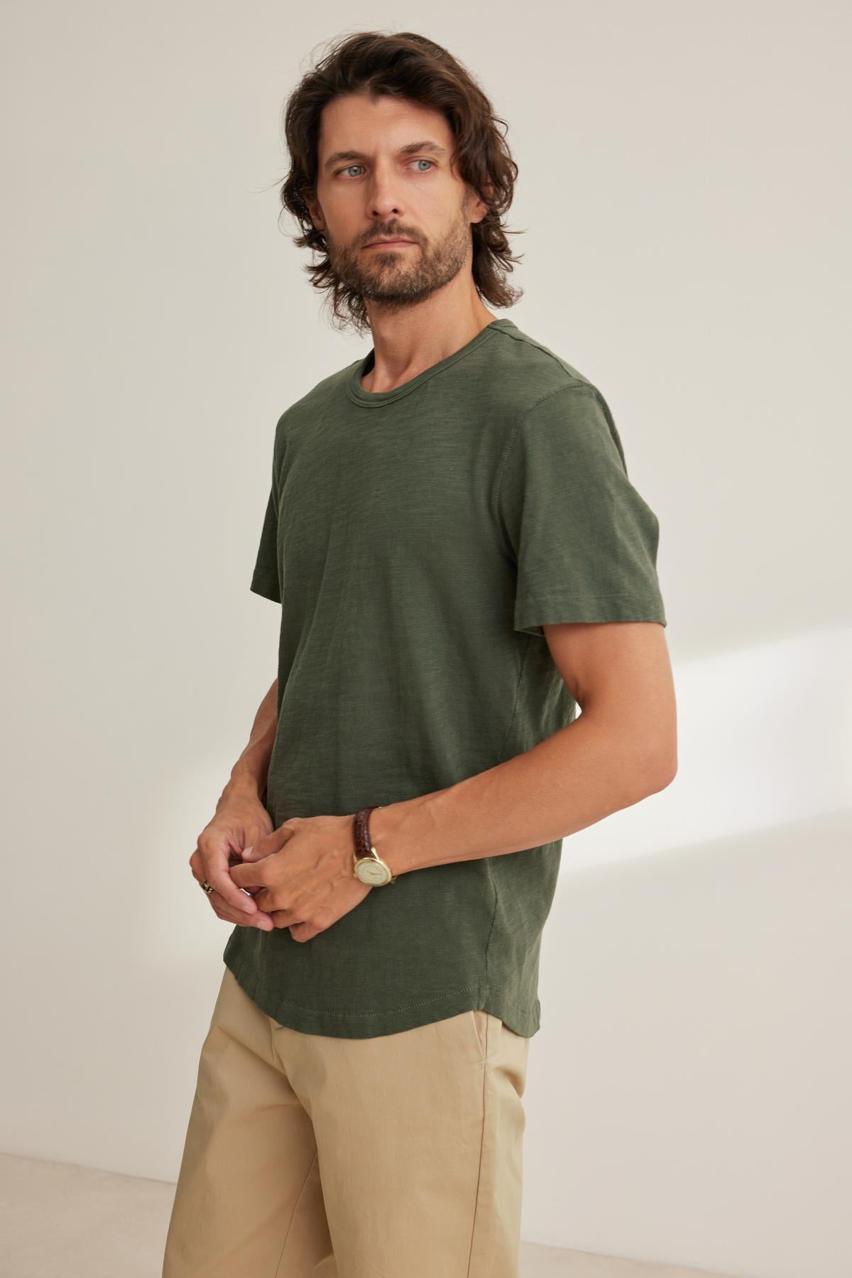   A man with medium-length hair wears a SPADER TEE by Velvet by Graham & Spencer, featuring a classic crew neckline and relaxed fit in green, paired with beige pants. Crafted from mid-weight Peruvian cotton, the outfit enhances his easygoing style against the plain background. 