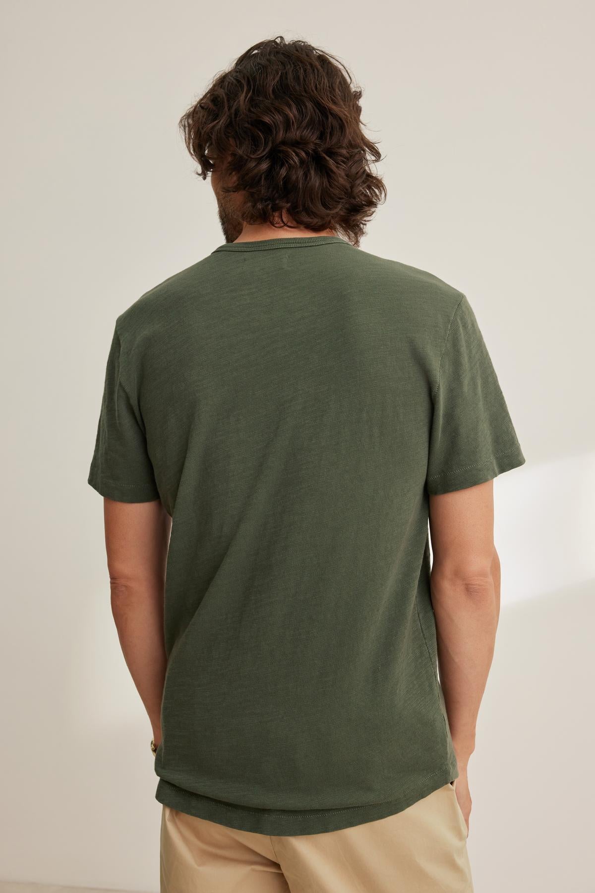   A person with medium-length hair faces away, wearing a Velvet by Graham & Spencer SPADER TEE in dark green with a classic crew neckline and beige pants. The relaxed fit of the mid-weight Peruvian cotton tee complements the softly lit room's ambiance. 