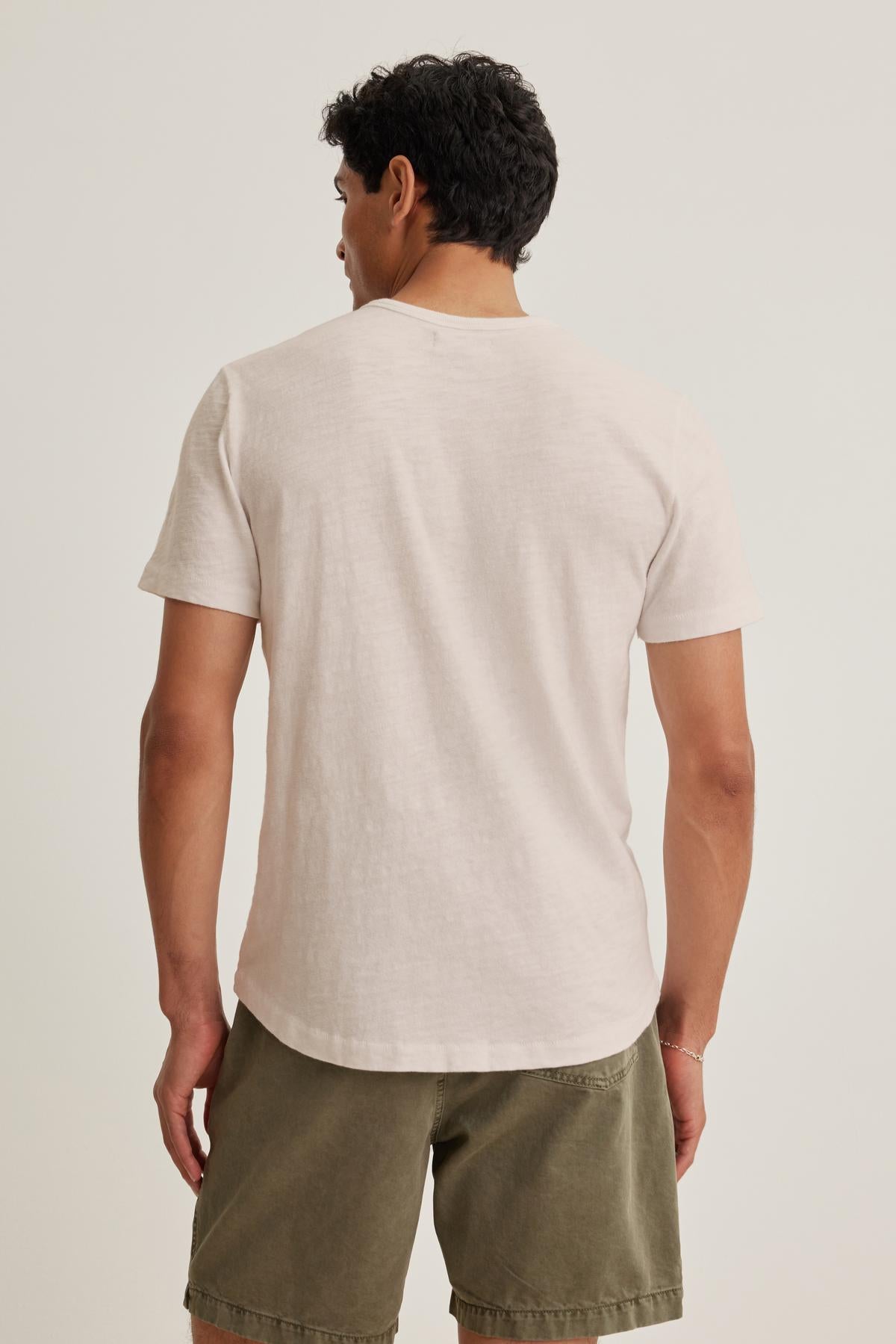   A laid-back man in the SPADER TEE by Velvet by Graham & Spencer, a relaxed-fit mid-weight Peruvian cotton white t-shirt, and olive shorts is seen from behind against a plain backdrop. 