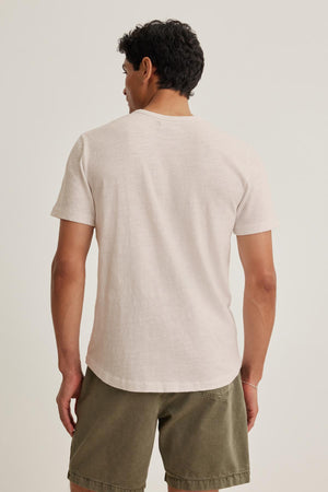 A laid-back man in the SPADER TEE by Velvet by Graham & Spencer, a relaxed-fit mid-weight Peruvian cotton white t-shirt, and olive shorts is seen from behind against a plain backdrop.