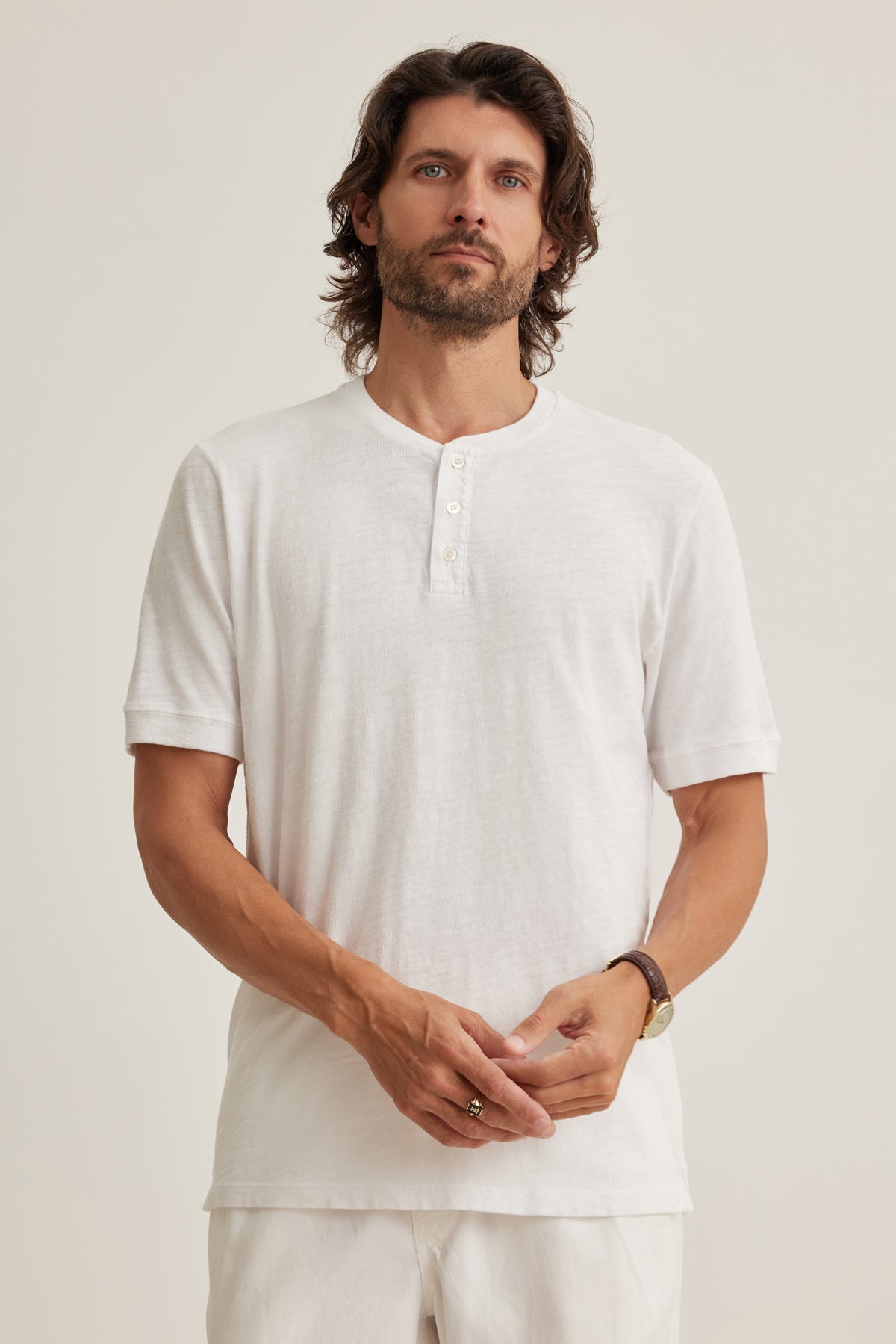 A man with wavy brown hair and a beard wears the Velvet by Graham & Spencer VITO HENLEY, a white short-sleeve Peruvian cotton shirt, with white pants, standing against a plain light background.-39161247006913