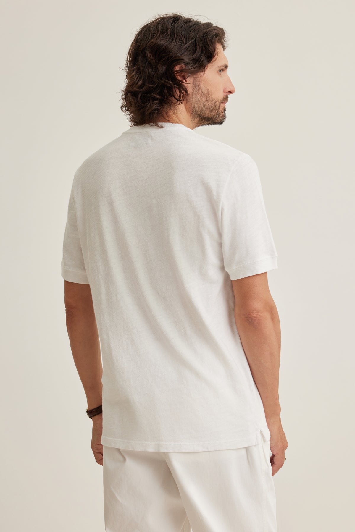 A man with shoulder-length dark hair stands with his back to the camera, wearing the Velvet by Graham & Spencer VITO HENLEY, a plain white Peruvian cotton short-sleeve t-shirt, and white pants against a neutral background.-39161247072449