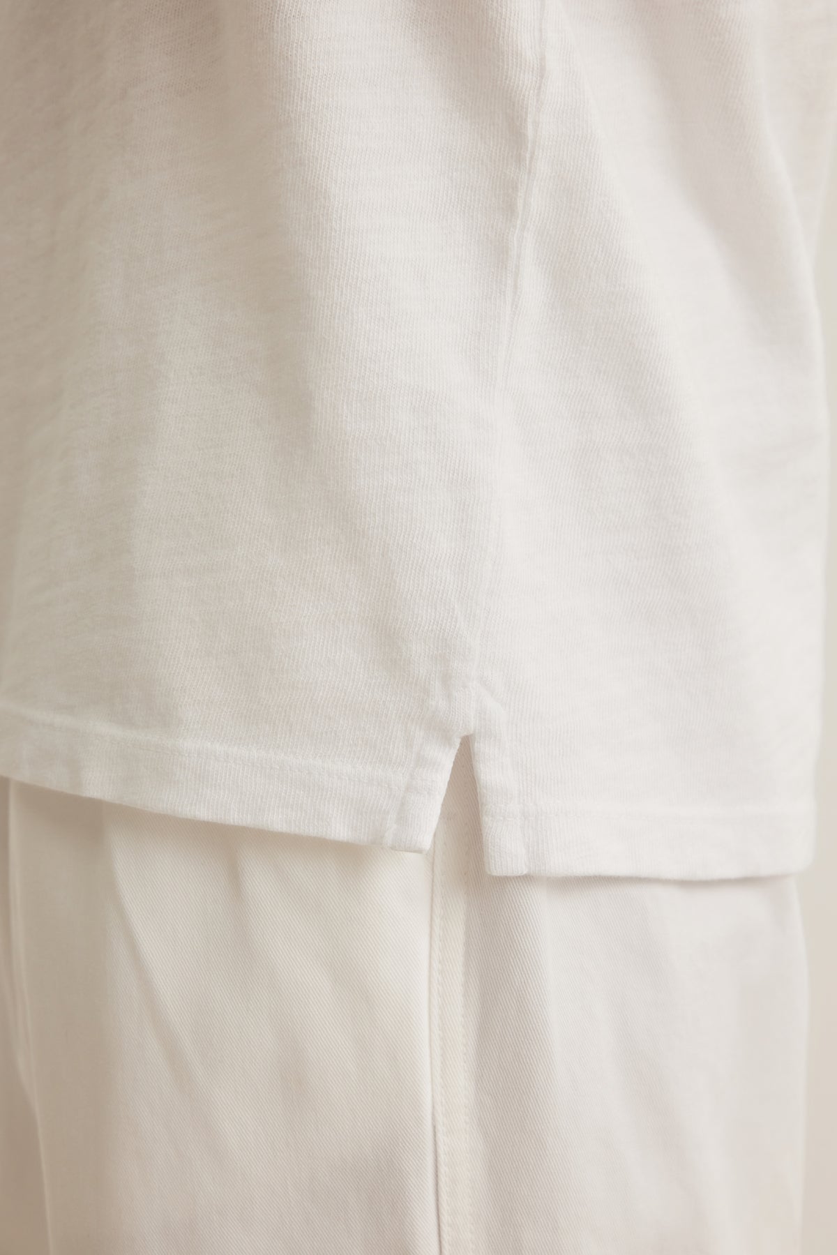 Close-up of the side seam and hem of the VITO HENLEY by Velvet by Graham & Spencer, showing the small slit at the bottom edge and its soft, textured feel in white Peruvian cotton worn over white pants.-39161247105217