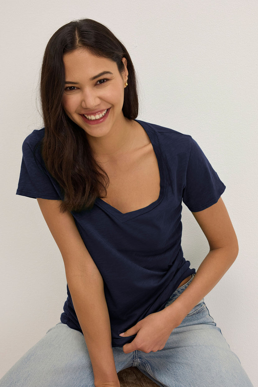 A person with long dark hair, wearing the LILITH TEE by Velvet by Graham & Spencer and jeans, sits and smiles against a plain background. The flattering fit of the cotton slub shirt enhances their relaxed and confident demeanor.