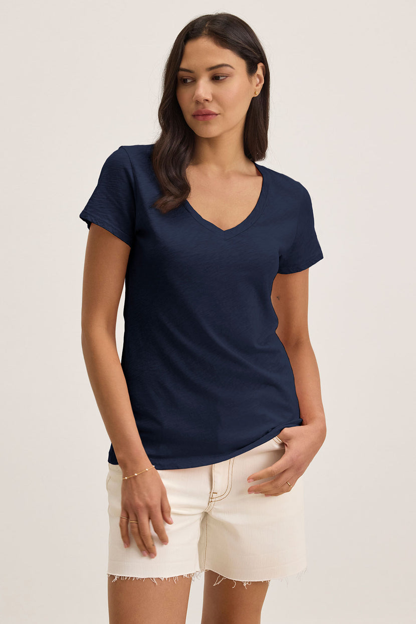 A woman wearing the luxurious Velvet by Graham & Spencer LILITH TEE and white shorts stands against a plain background, glancing to her left.