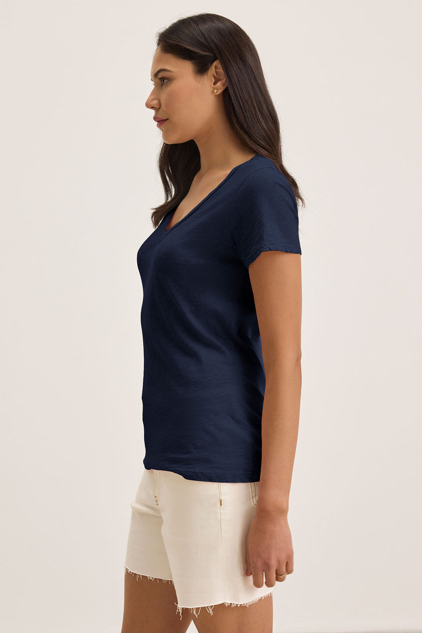 A woman with long dark hair, wearing Velvet by Graham & Spencer's LILITH TEE in navy and beige cotton slub shorts, stands sideways against a plain background.