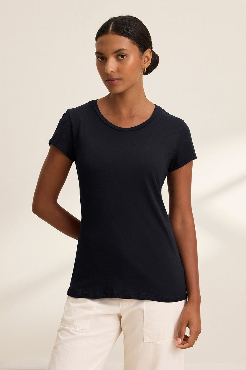 A person stands against a neutral background wearing the ODELIA TEE by Velvet by Graham & Spencer, a black premium cotton short-sleeve t-shirt, paired with light-colored pants and one arm behind their back.
