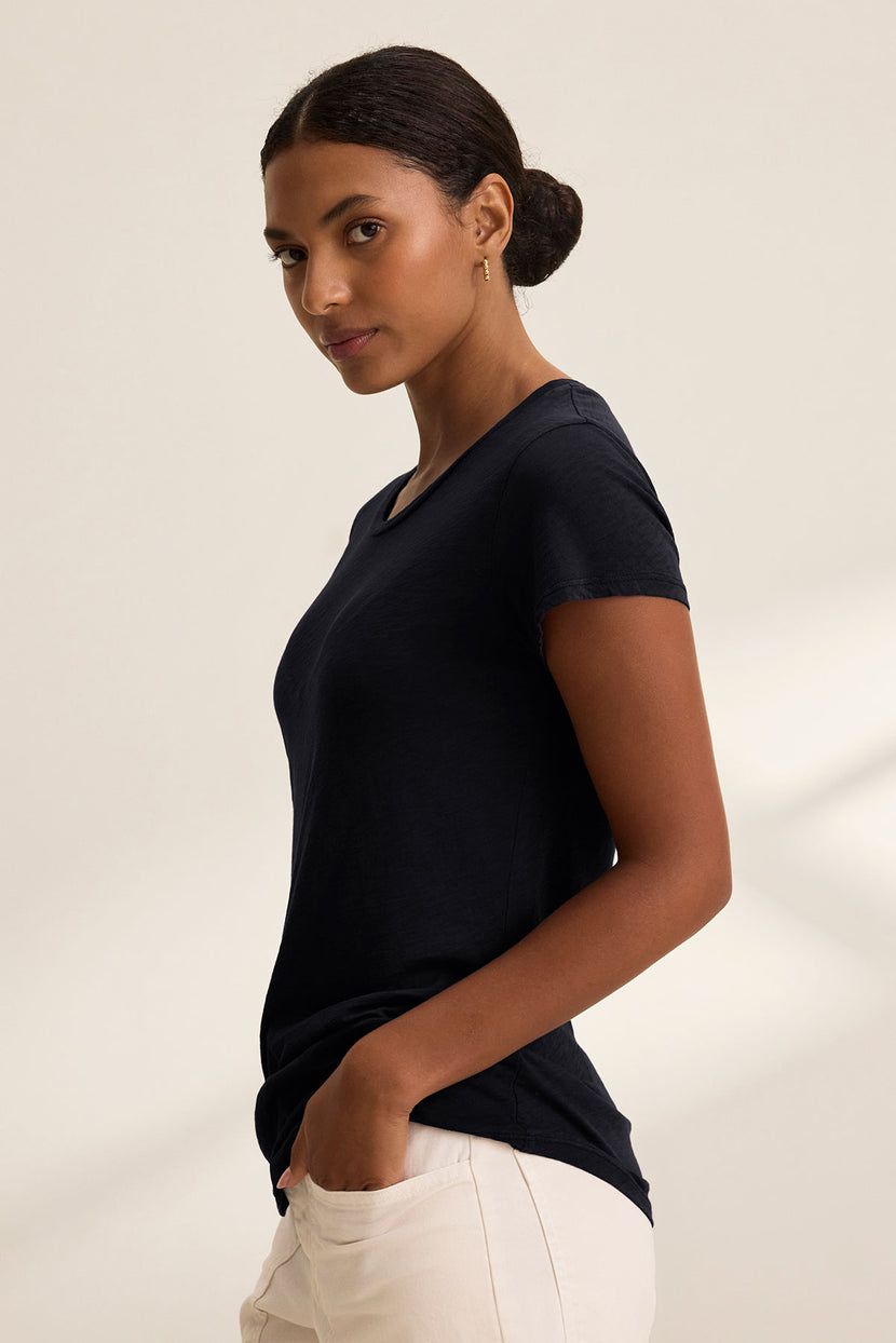 A woman in profile, hand in pocket, wears the ODELIA TEE by Velvet by Graham & Spencer—a premium cotton black short-sleeve shirt—paired with light-colored pants against a plain background.