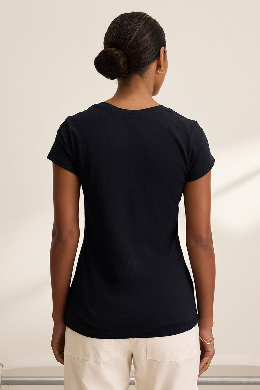 A woman with dark hair in a bun wears the ODELIA TEE by Velvet by Graham & Spencer, a black premium cotton short-sleeve t-shirt, paired with light pants and shown from the back against a neutral background.