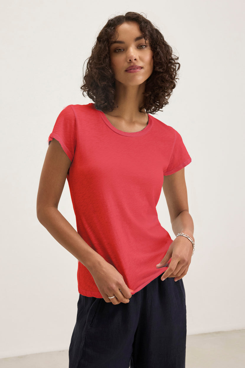 A person in a red ODELIA TEE by Velvet by Graham & Spencer, featuring a classic crew neckline and cozy cotton slub fabric, stands against a plain white background in dark pants, showcasing timeless style and comfort.