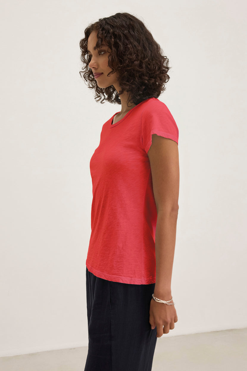 Against a plain white background, a person with curly hair is in profile, highlighting the Velvet by Graham & Spencer ODELIA TEE—a classic red crew-neck t-shirt made of cotton slub fabric—paired stylishly with dark pants.