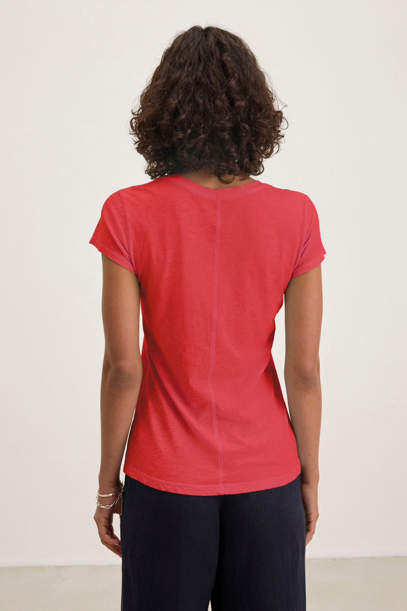 A curly-haired person faces away from the camera, wearing the ODELIA TEE from Velvet by Graham & Spencer—a red cotton slub short-sleeve essential—and dark pants against a plain white background.