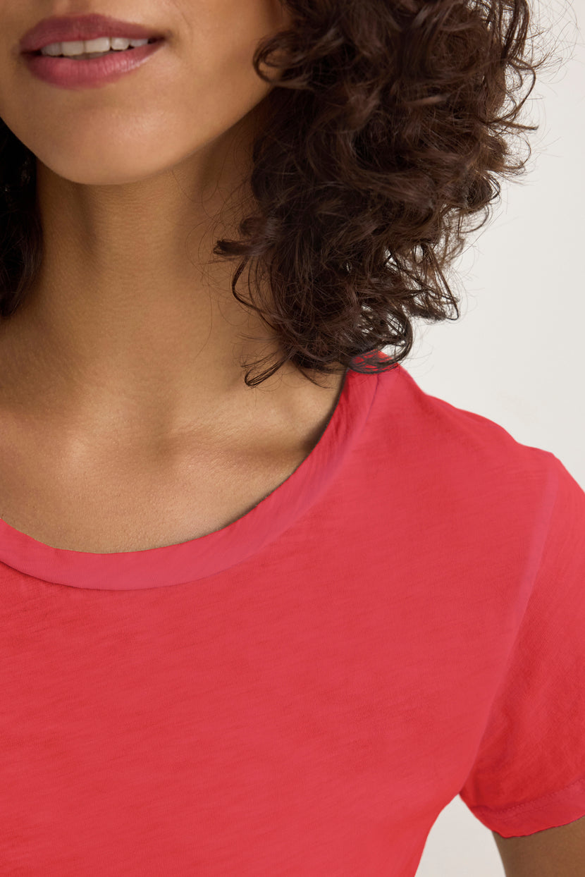 Partial view of a person with curly hair in the ODELIA TEE by Velvet by Graham & Spencer, a red cotton slub fabric shirt. The glimpse reveals part of the face and neck, accentuated by a classic crew neckline—a true wardrobe essential.