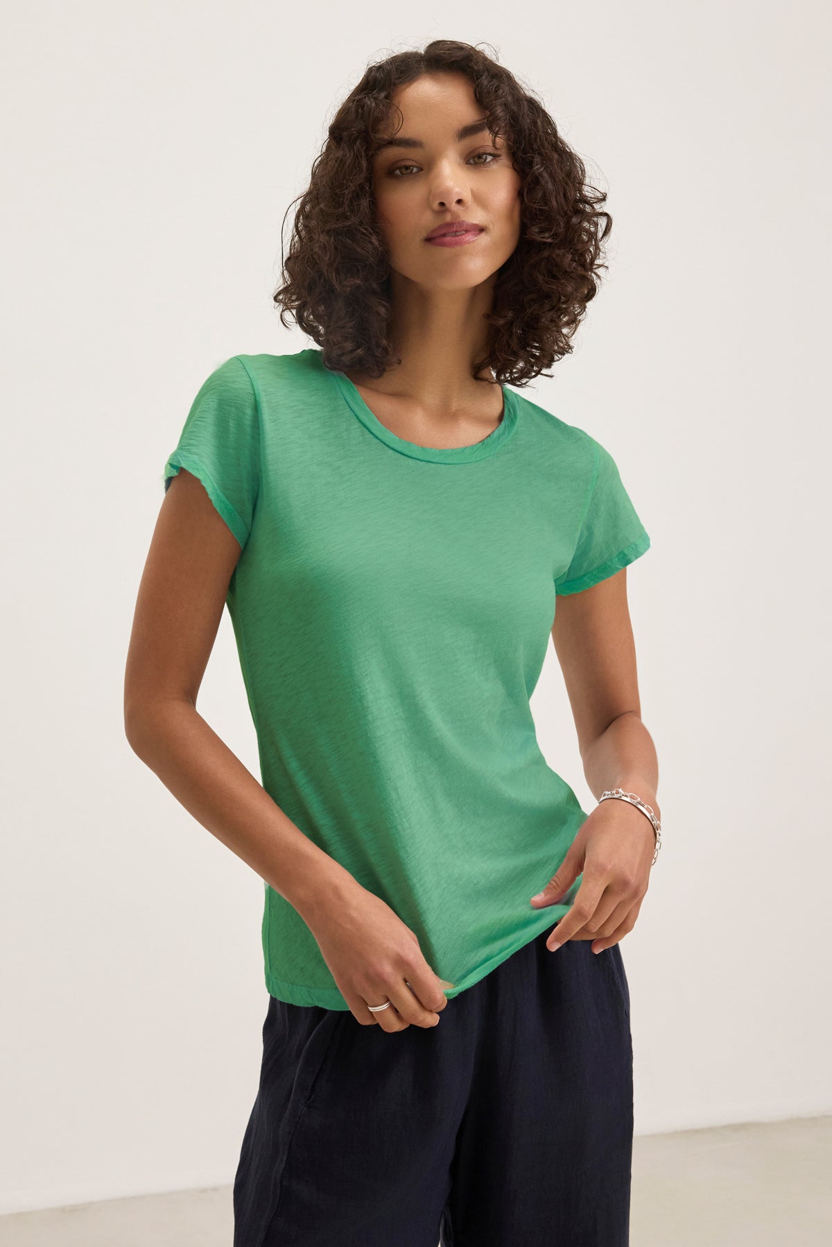 A woman with curly hair wears the Velvet by Graham & Spencer ODELIA TEE in green and dark pants, standing against a plain light background.-39395676160193