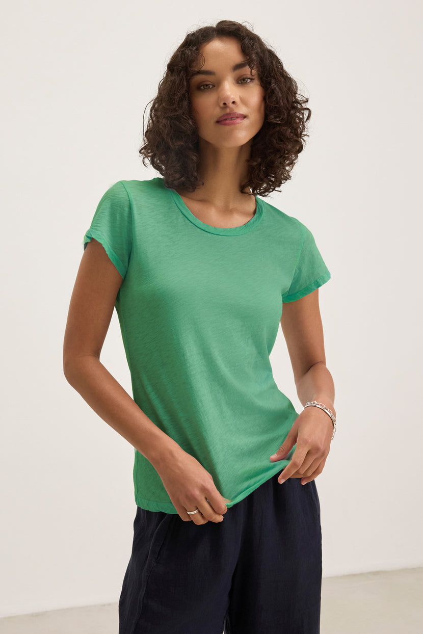 A woman with curly hair wears the Velvet by Graham & Spencer ODELIA TEE in green and dark pants, standing against a plain light background.