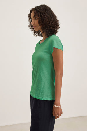 A woman with curly hair wears the ODELIA TEE by Velvet by Graham & Spencer, a green short-sleeve crew neck, paired with dark pants, standing in profile against a plain light background.