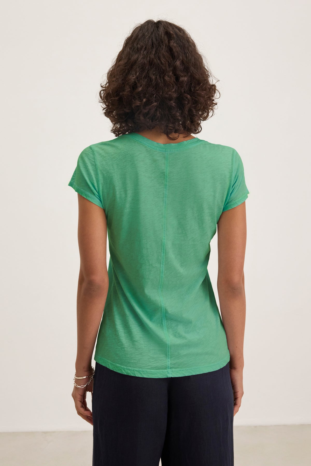 Someone with curly hair is seen from behind, wearing the Velvet by Graham & Spencer ODELIA TEE in green—a short-sleeve, crew neck t-shirt—paired with dark pants and standing against a plain background.-39395676094657