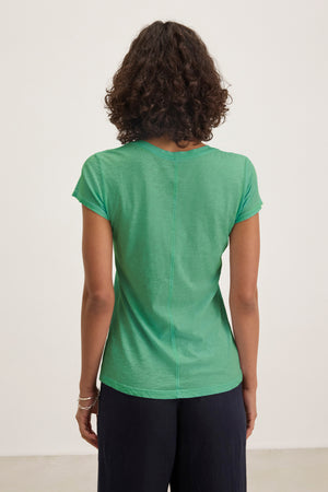 Someone with curly hair is seen from behind, wearing the Velvet by Graham & Spencer ODELIA TEE in green—a short-sleeve, crew neck t-shirt—paired with dark pants and standing against a plain background.